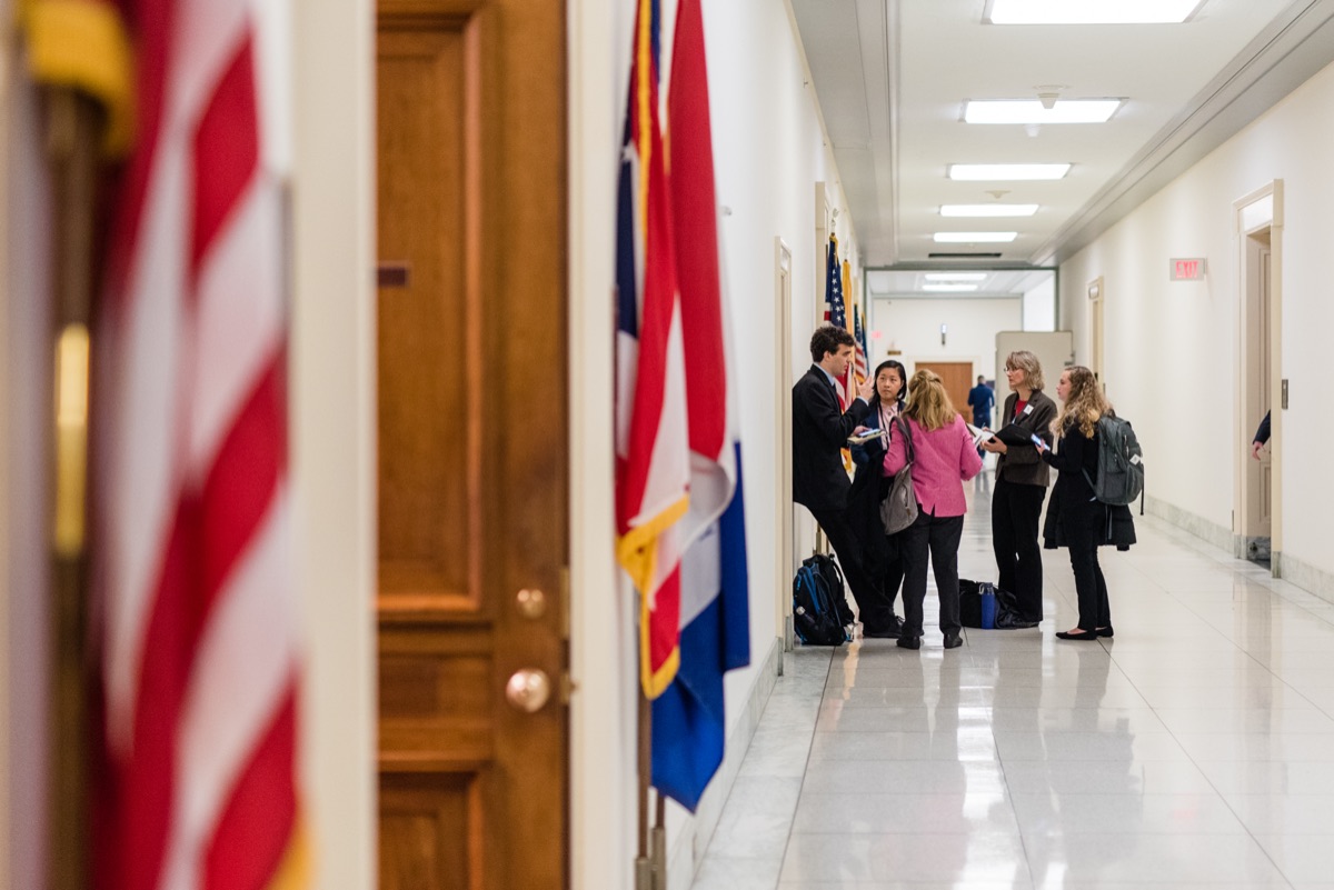 A lobby team gathers down a congressional hallway, framed by flags